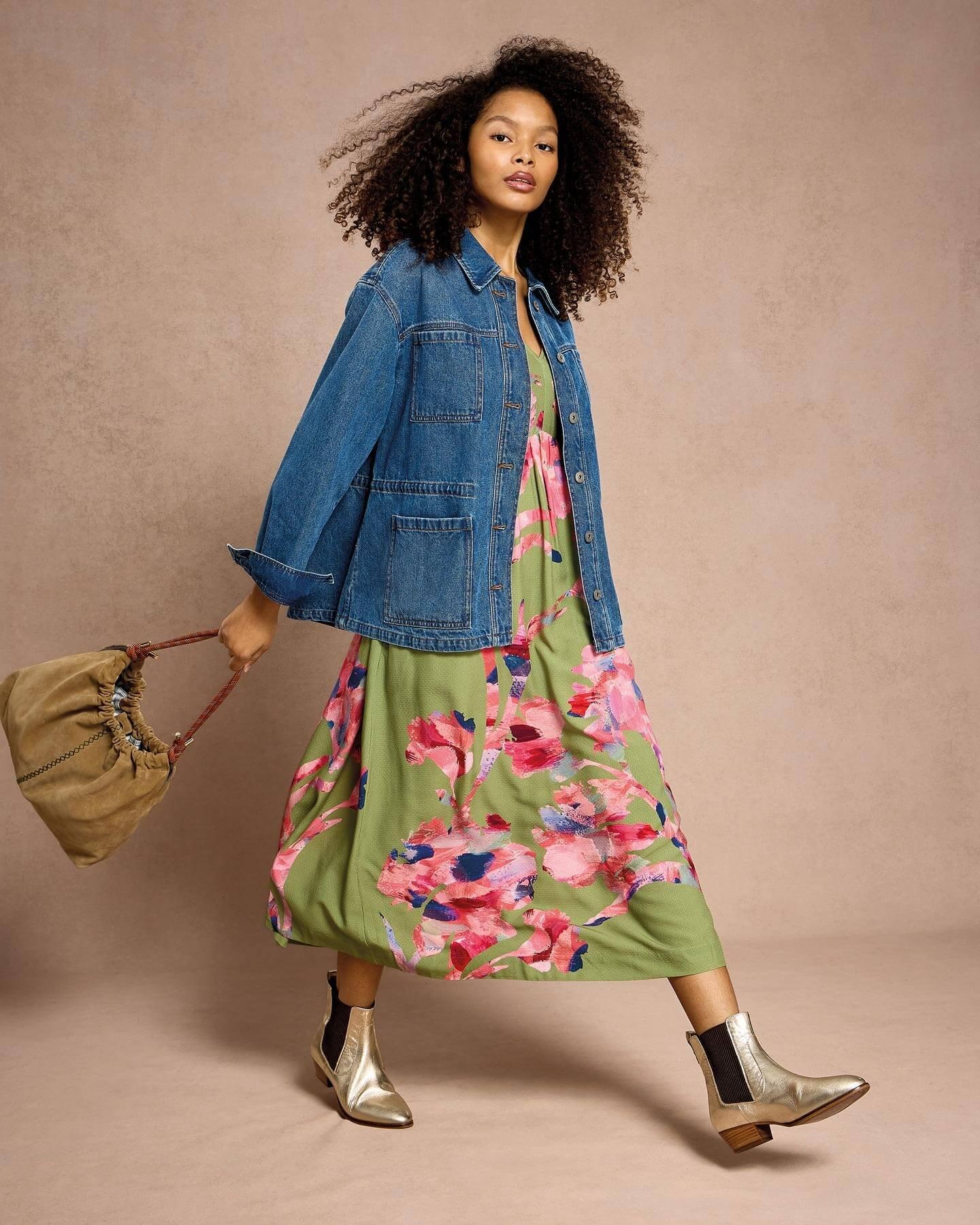 a woman walking in front of a pink background wearing metallic boots, a green floral printed dress, blue denim jacket and a brown suede shoulder bag