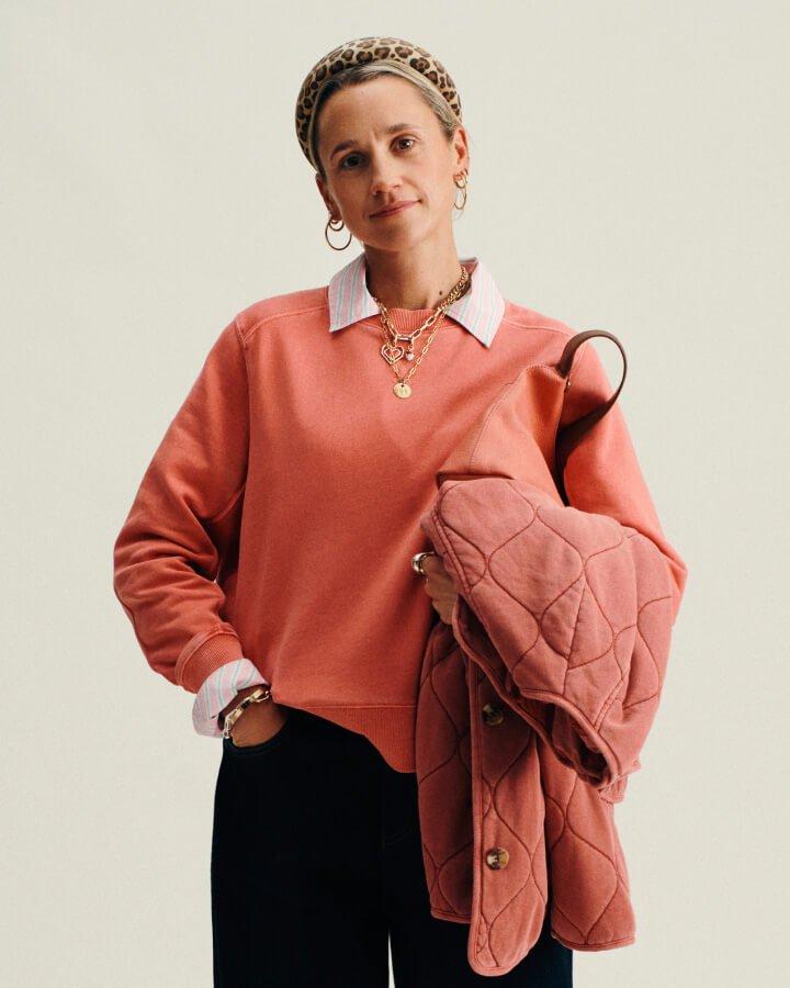 a woman stood up wearing a pair of black trousers, stripey pink shirt with an orange sweatshirt layered over the top. In her left hand she's holding a coat and bag.