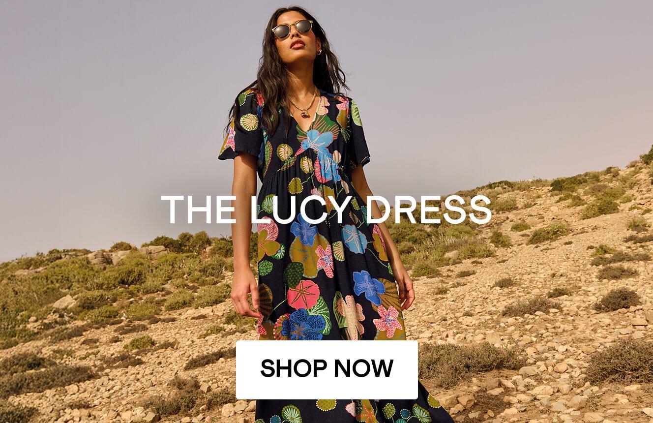 A woman wearing a black dress with geometric patterns in the desert, with the text "The Lucy Dress", Click here to shop more Lucy dresses. 