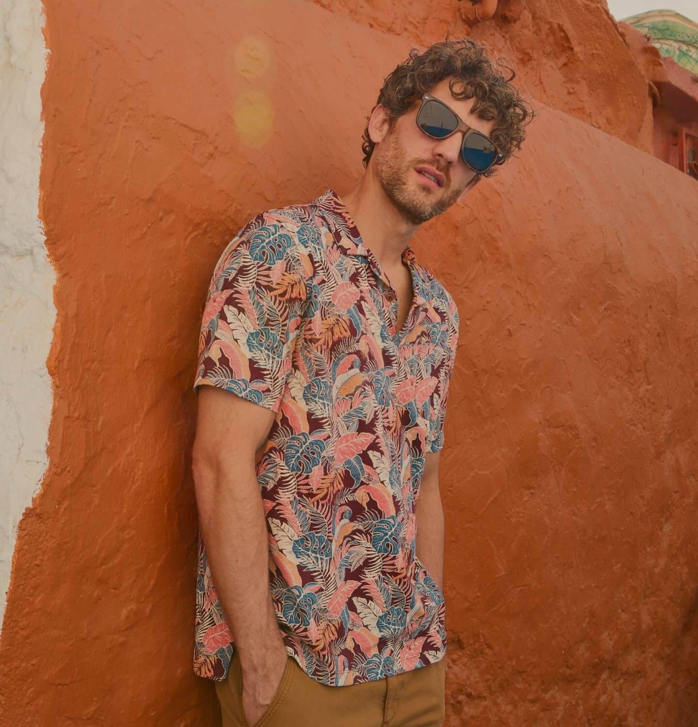 Man in sunglasses wearing a patterned short sleeved shirt and leaning against an orange wall