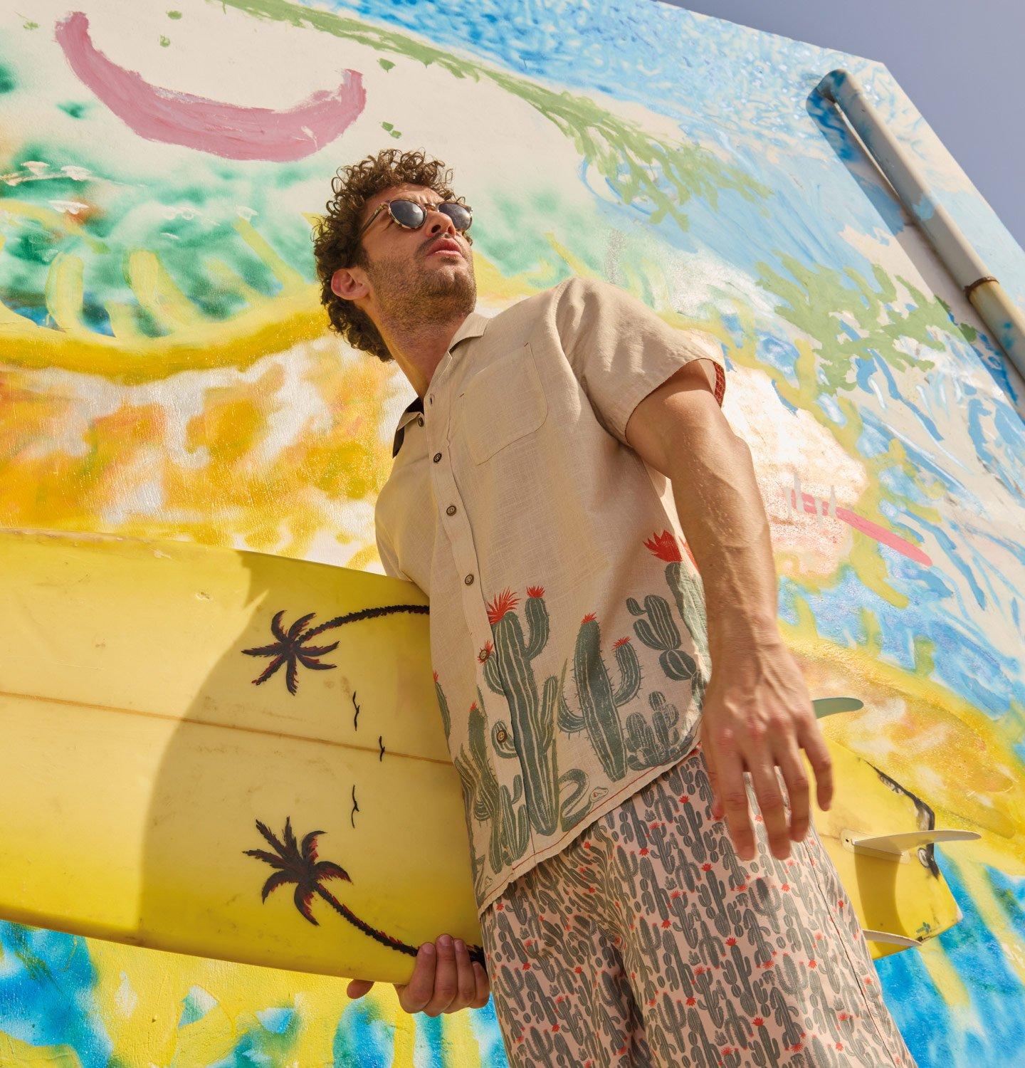 A male model holding a surfboard in front of a colorful mural, wearing the Hayle Cactus Printed Shirt from White Stuff.