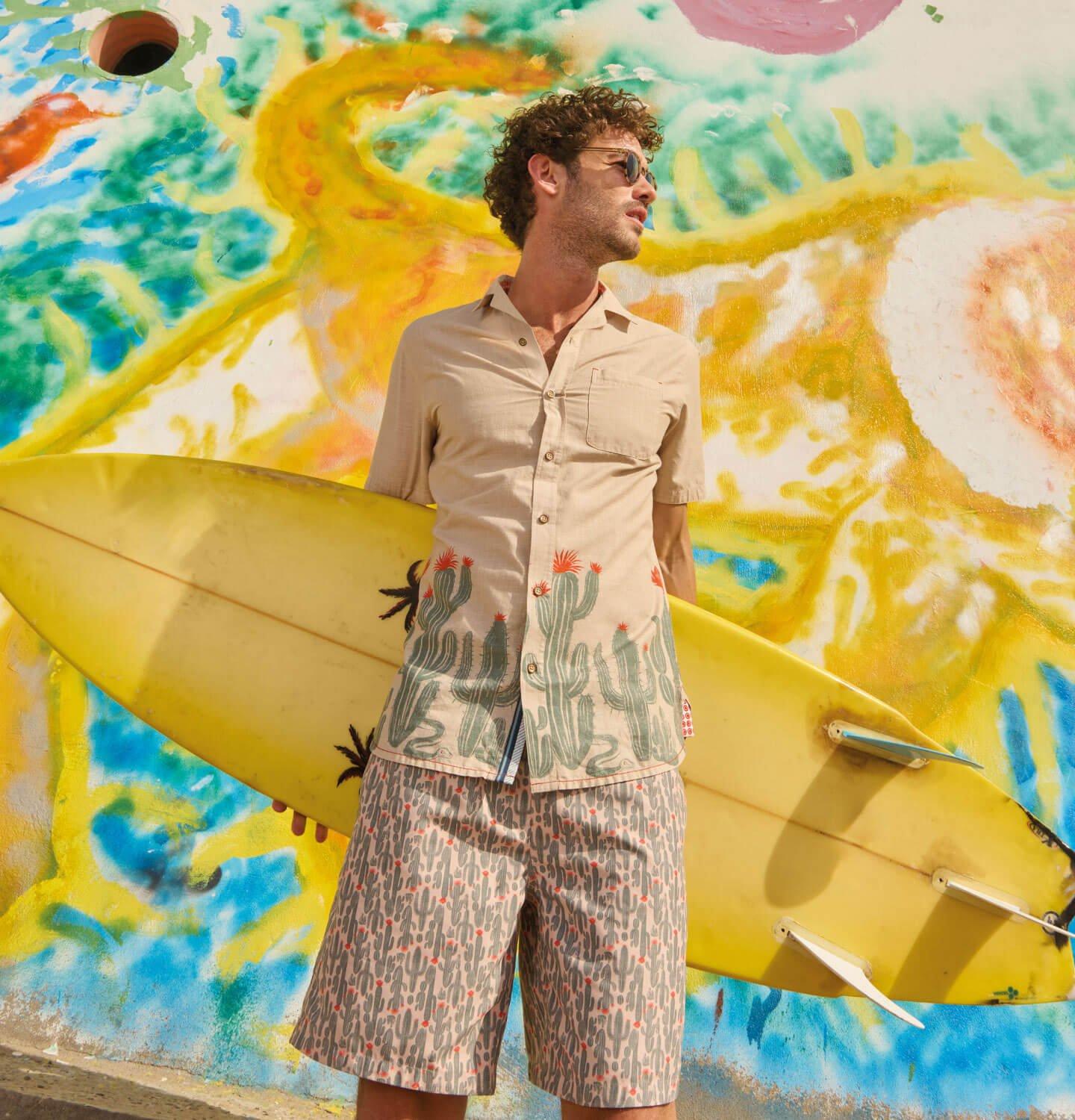 A male model holding a surfboard in front of a colorful mural, wearing the Hayle Cactus Printed Shirt from White Stuff.