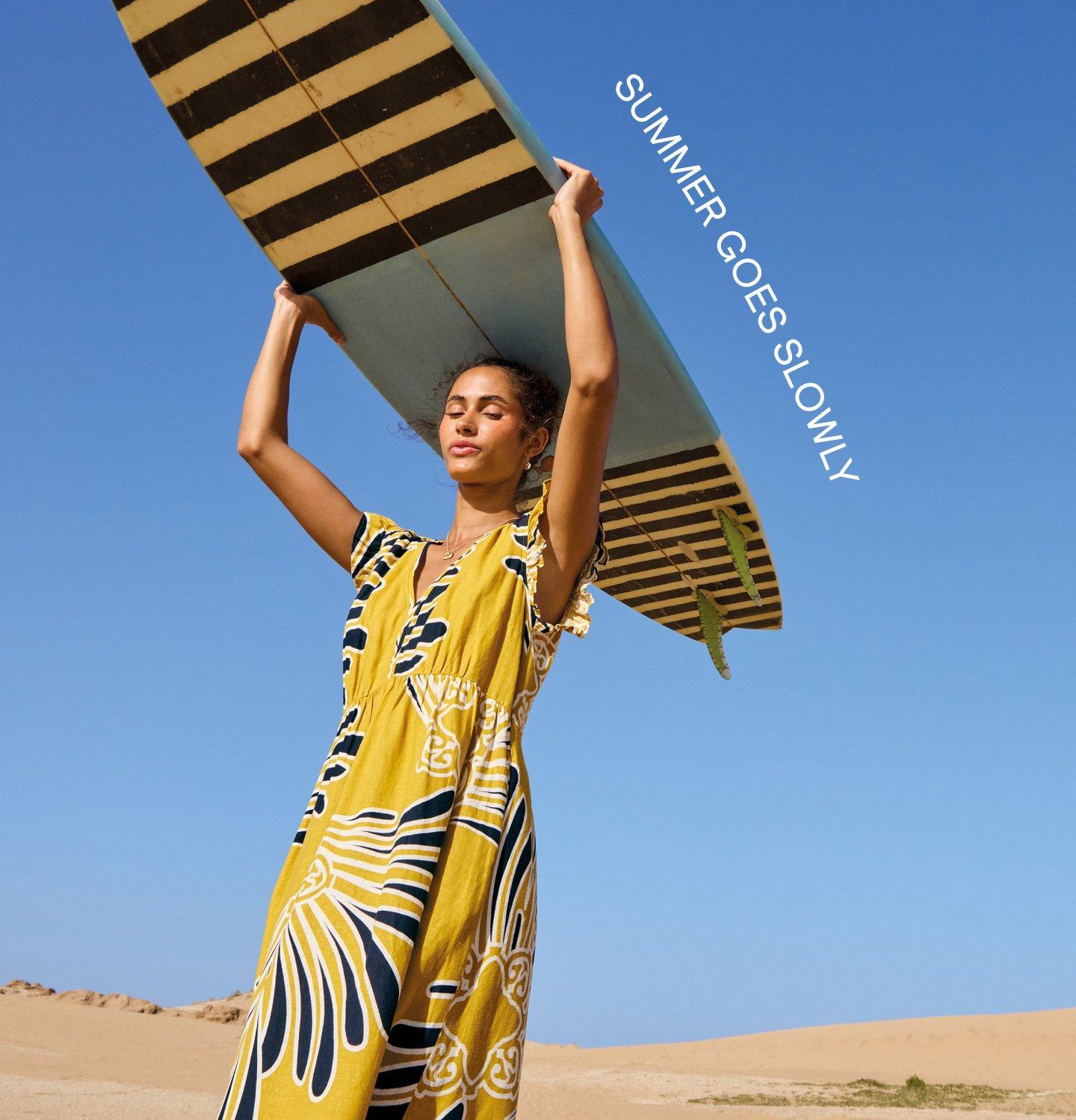 A female model wearing the yellow Lorna Maxi Linen Blend Dress is holding a surfboard above her head. The text on the image reads Summer Goes Slowly.