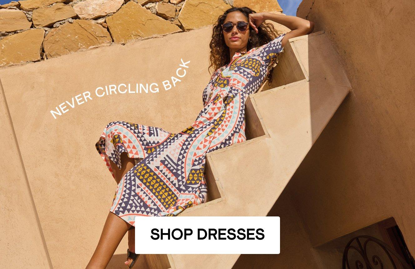 A woman is sitting on stairs wearing a dress with a geometric pattern, the setting is the desert, there is writing on the image saying "Never circling back". Click here to shop dresses.
